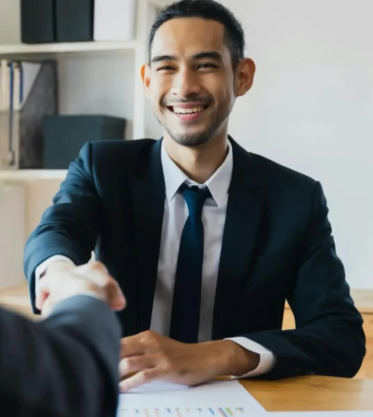 Smiling businessman in a suit shaking hands across a desk.