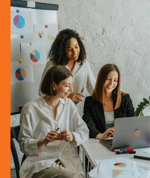 Three women collaborating in front of a laptop with charts in the background.