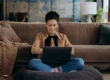 A smiling woman sitting cross-legged on a cushion in front of a laptop, appearing focused and relaxed, with a cozy living room in the background.