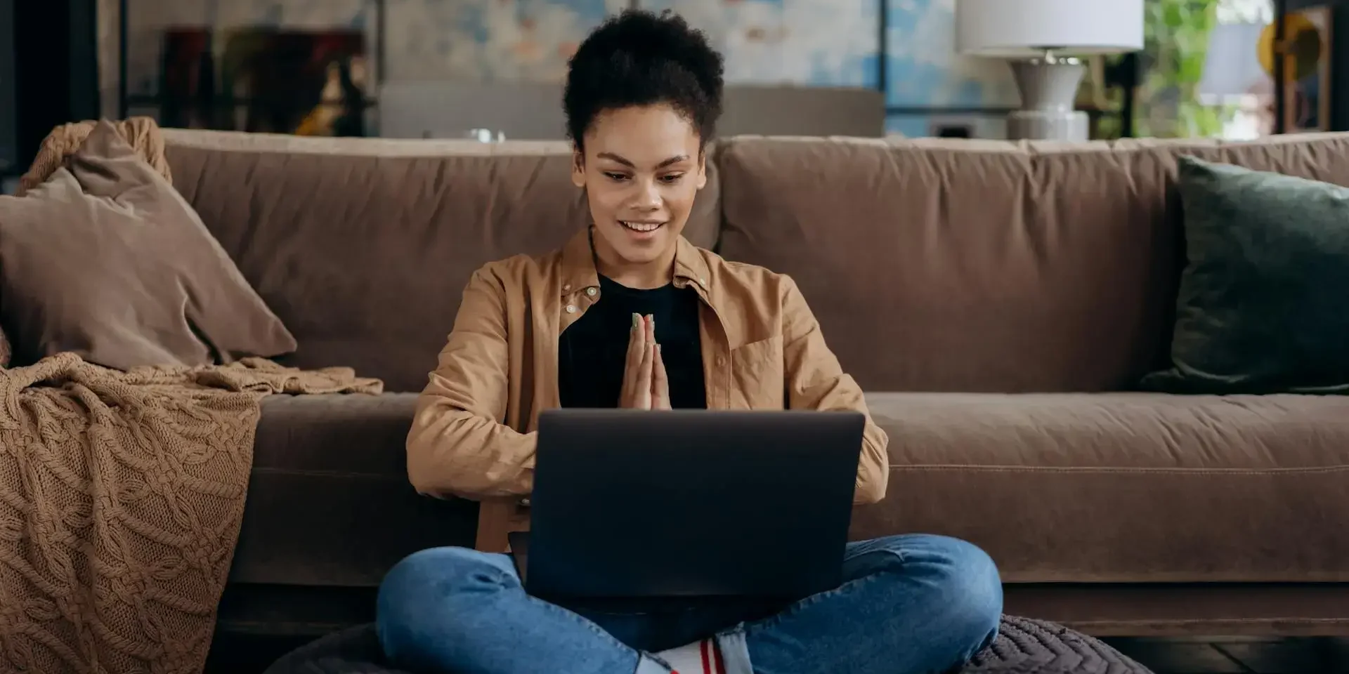A smiling woman sitting cross-legged on a cushion in front of a laptop, appearing focused and relaxed, with a cozy living room in the background.