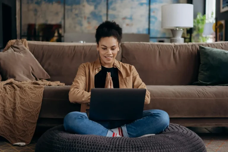 Woman Using Laptop for Work in a Relaxed Setting A smiling woman sitting cross-legged on a cushion in front of a laptop, appearing focused and relaxed, with a cozy living room in the background.